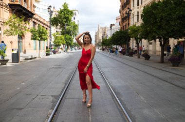 Kırmızı ipek elbiseli güzel bir kadın Seville 'de bir caddede tramvay raylarının arasında duruyor. Model fotoğraf çekimi için poz veriyor. Moda ve güzellik konsepti.