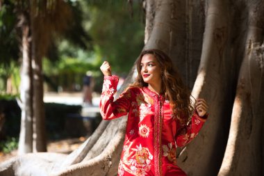 Güzel bir Faslı kadın geleneksel Fas kırmızısı, altın ve gümüş işlemeli bir elbise giyiyor. Kadın bir ağacın gövdesinin yanındaki parkta. Şehre seyahatin tadını çıkarıyor..
