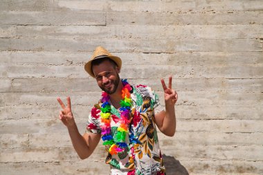 Havai çiçekli gömlek, şapka ve renkli çiçekli kolye giyen yakışıklı bir genç. Adam yüz ve vücut ifadeleriyle farklı jestler yapıyor. Arkaplan gri duvarı.