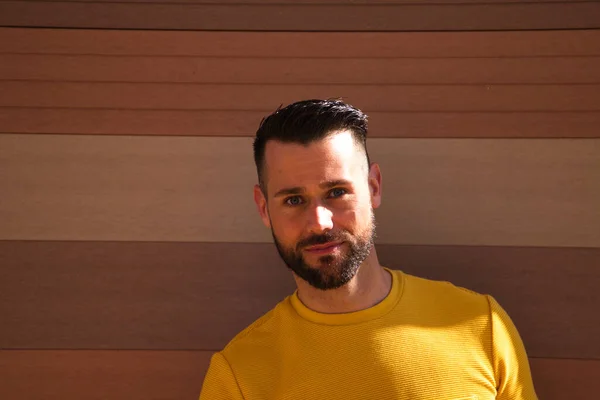 A young, handsome, blue-eyed, bearded man with a yellow T-shirt holds a hat in his hands. In the background a wall in shades of brown. The man smiles happily looking forward to the holidays.
