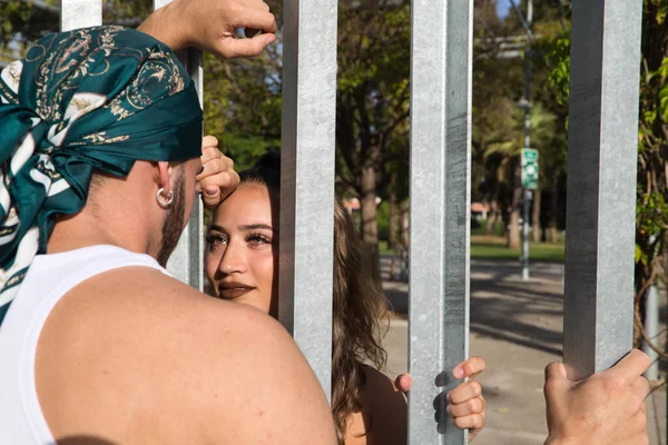 Portrait of a couple in love separated by a fence. They look at each other emotionally with sadness and sorrow because they are each on one side of the fence. The young couple are dressed fashionably.