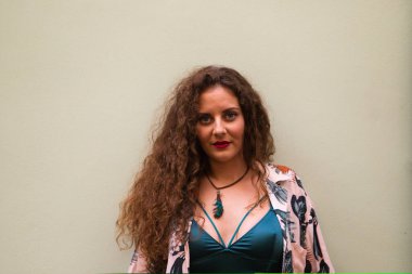 Kıvırcık saçlı ve mavi gözlü genç sarışın kadın bej bir duvarın önünde değişik jestler ve ifadeler yapıyor. İfadeleri kavra.