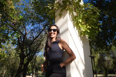 Güzel, esmer, siyah güneş gözlüklü İspanyol kadın. Kadın parkta bir parkın tepesinde yatıyor. Kadın mutlu bir şekilde gülümsüyor ve siyah kıyafetler giyiyor..