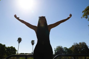 Güzel esmer İspanyol kadın kollarını gökyüzüne kaldırıyor. Arka planda bir göl var ve ışık huzmeleri arkadan geliyor. Kadın zarif siyah elbiseler giyiyor..