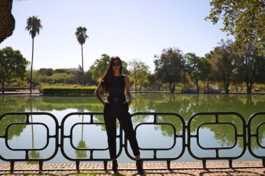 Güzel esmer İspanyol kadın parktaki bir gölün korkuluğuna yaslanıyor. Kadın zarif siyah elbiseler giymiş ve güneş gözlüğü takıyor..