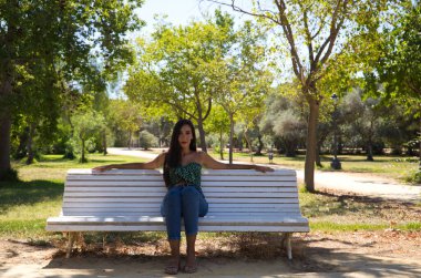 Siyah saçlı güzel İspanyol kadın parkta beyaz bir bankta oturuyor. O yalnız ve düşünceli. İfadeleri ve jestleri kavra.