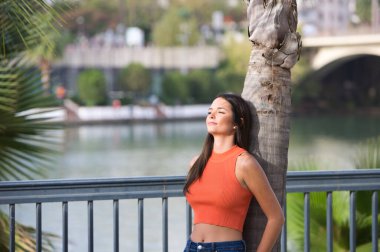 Young and beautiful woman leaning on the trunk of a palm tree. Woman is happy and relaxed and enjoying the sunny day in seville, spain. The woman is wearing jeans and orange top.