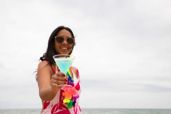 A young, beautiful Latina woman is holding a glass with a blue cocktail in it. The woman is on the beach wearing sunglasses. In the background you can see the sea. Holiday and travel concept.