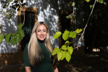 Yeşil elbiseli güzel sarışın kadın bir ışık huzmesinin geçtiği ağacın dallarının yanında duruyor. Doğayla temas.