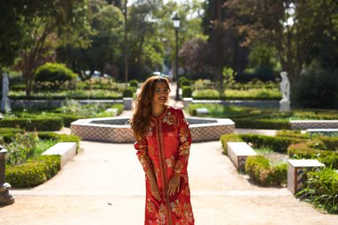 Güzel bir kadın geleneksel bir Fas elbisesi giyer. Kırmızı ve gümüş işlemeli. Kız Fas tarzı bir bahçede, fıskiyeli ve yemyeşil bir yerde düğün fotoğrafları için poz veriyor..