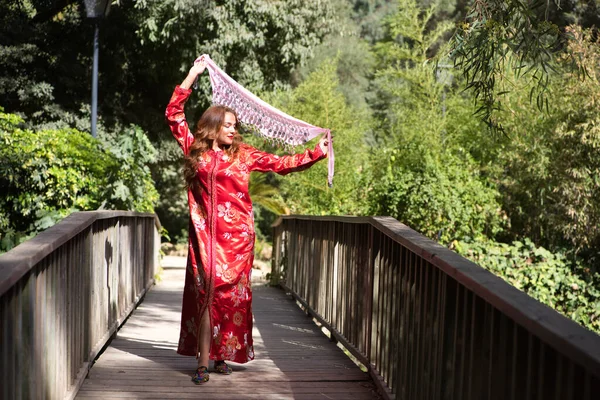 A beautiful Moroccan woman is wearing a traditional Moroccan red dress with gold and silver embroidery. The woman is dancing with a pink scarf on a wooden bridge among the vegetation.