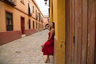 Young, blonde, beautiful woman in a red dress is visiting seville. The woman poses for the camera very elegant and like a model in the typical streets of the city. Holidays and travels