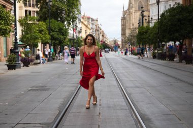 Büyük bir şehirde tramvay rayları arasında koşan kırmızı ipek elbiseli güzel bir kadın. Manken tek eliyle elbisesini tutuyor. Moda ve güzellik konsepti.