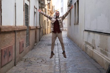 Sakallı ve gözlüklü yakışıklı genç bir adam dar bir sokakta sevinçten zıplıyor. Mutluluk ve neşe kavramı.