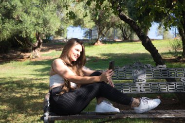 Genç, güzel, sarışın, mavi gözlü bir kadın bankta oturmuş cep telefonuyla selfie çekerken ve sosyal medyayı kontrol ederken mutlu..