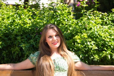 Parktaki bankta oturan mavi gözlü ve kısa elbiseli güzel sarışın kadının portresi. Kadın bacak bacak üstüne atmış dinleniyor ve mutlu..