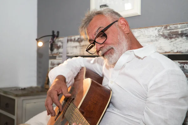 Senior man with white beard sitting on bed playing guitar. Man is in pajamas and has glasses. Senior and grandparents concept.