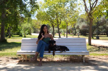 Güzel siyah saçlı kadın parktaki beyaz bankta doberman köpeğiyle oturuyor. Kadın köpeğiyle vakit geçirmekten ve birlikte dinlenmekten hoşlanıyor. Evcil hayvanlar ve hayvanlar konsepti.