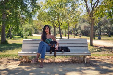 Güzel siyah saçlı kadın parktaki beyaz bankta doberman köpeğiyle oturuyor. Kadın köpeğiyle vakit geçirmekten ve birlikte dinlenmekten hoşlanıyor. Evcil hayvanlar ve hayvanlar konsepti.