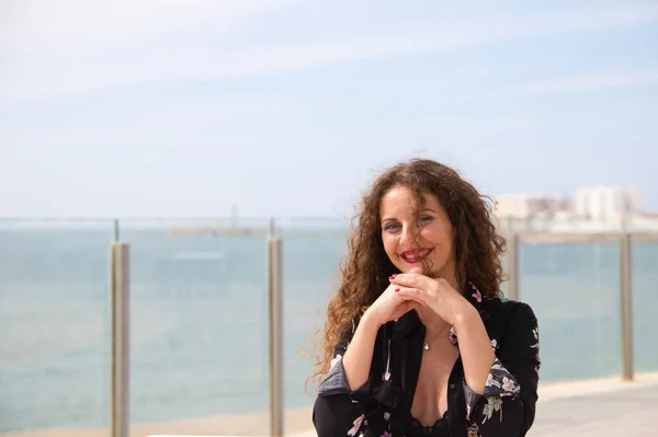 Beautiful blonde woman with curly hair and blue eyes is sitting on the promenade. In the background you can see the sea. The woman looks at camera and makes different expressions, happy, sad.
