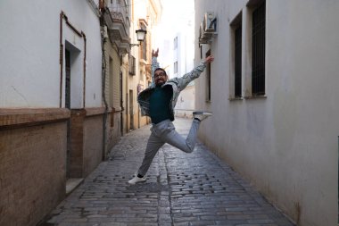 Sakallı ve gözlüklü yakışıklı genç bir adam dar bir sokakta sevinçten zıplıyor. Mutluluk ve neşe kavramı.