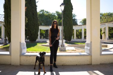 Doberman köpeğiyle güzel esmer kadın. Kadın siyah kıyafetler giyiyor ve köpeği zincirle tutuyor. Evcil hayvanlar ve hayvanlar konsepti.