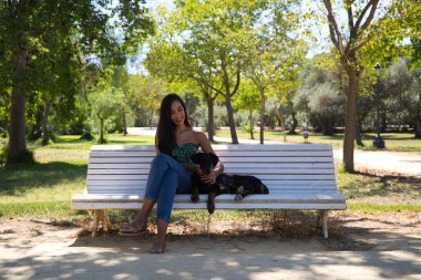 Güzel siyah saçlı kadın parktaki beyaz bankta doberman köpeğiyle oturuyor. Kadın köpeğiyle vakit geçirmekten ve birlikte dinlenmekten hoşlanıyor. Evcil hayvanlar ve hayvanlar konsepti.