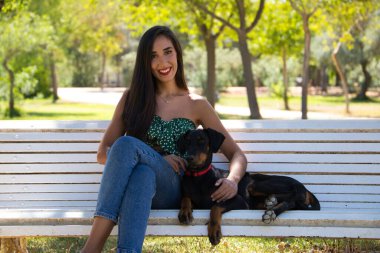 Güzel siyah saçlı kadın parktaki beyaz bankta doberman köpeğiyle oturuyor. Kadın köpeğiyle vakit geçirmekten ve birlikte dinlenmekten hoşlanıyor. Evcil hayvanlar ve hayvanlar konsepti.