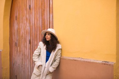 Young and beautiful Hispanic brunette woman with curly hair with hat and coat for the cold leaning on wall of a house in seville. Travel and holiday concept.