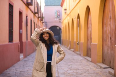 Young and beautiful Hispanic brunette woman with curly hair wearing a hat and coat for the cold walking in the city of seville while making different expressions and having fun.