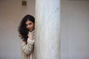 Young and beautiful brunette woman with curly hair and a woollen coat for the winter, leaning on a marble column while making different facial and body expressions.