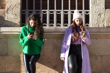 Two young and beautiful Spanish women are consulting the mobile phone in a street of a big city. The women are having fun while consulting the mobile phone.