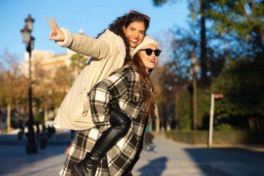 two young, beautiful Spanish women who are friends have fun playing on top of each other while they are walking and enjoying the sunny winter day. The women are wearing winter clothes.