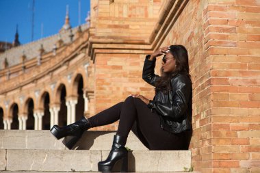 young and beautiful black latin woman wearing black clothes and sunglasses is sitting on the steps of a square in seville, spain. The photo is taken in profile and the girl is doing different poses.