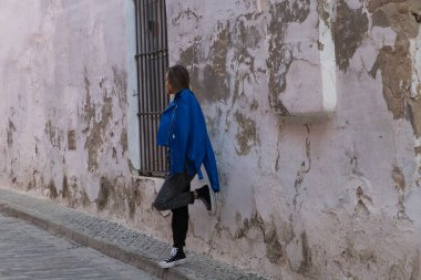 Young blonde woman dressed in black t-shirt and jeans leaning against an old wall and wearing blue jacket in . The young woman makes different gestures and body expressions.