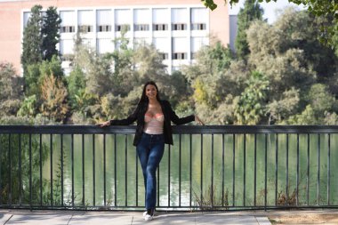 Beautiful young woman leaning on the river railing in Seville, Spain. In the background you can see the river and part of the city. The woman is dressed casual.