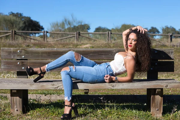 スペイン人 ブルネット 巻き毛の女性が森につながる道のベンチに横たわっています 彼女はジーンズと白い上に身を包み 晴れた日を楽しんでいます — ストック写真