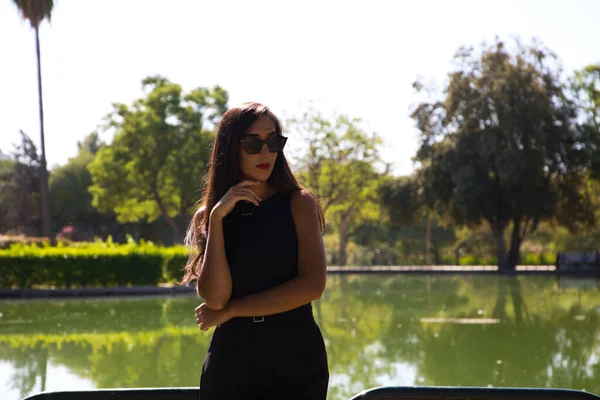 美丽的黑发西班牙女人靠在公园湖畔的栏杆上 这个女人穿着雅致的黑色衣服 戴着太阳镜 — 图库照片