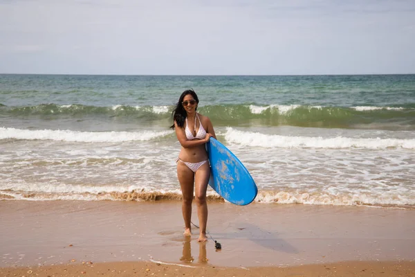美しい若いラテン語の女性ビキニは青いサーフボードで海から出てくる その女性は浜辺にいる 休日と夏のコンセプト — ストック写真
