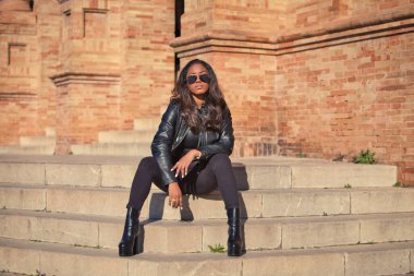 young and beautiful black latin woman wearing black clothes and sunglasses is sitting on the steps of the most important square in the city of seville, spain. The photo is taken from the front.