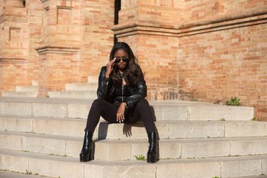 young and beautiful black latin woman wearing black clothes and sunglasses is sitting on the steps of the most important square in the city of seville, spain. The photo is taken from the front.