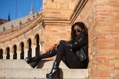 young and beautiful black latin woman wearing black clothes and sunglasses is sitting on the steps of a square in seville, spain. The photo is taken in profile and the girl is doing different poses.