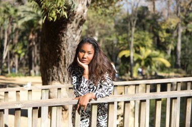 beautiful young black latin woman leaning on the railing of a wooden bridge in a park in seville looking at the camera with her face in her hands.