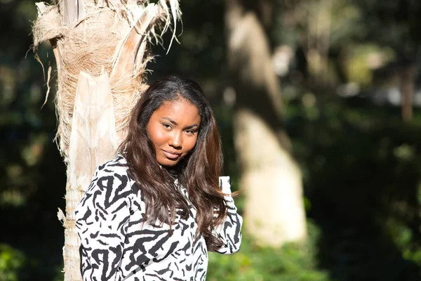 beautiful young black latin woman wearing a black and white dress and leaning on the trunk of a tropical palm tree while looking at the camera. Holiday and travel concept.