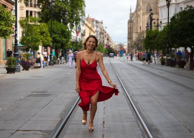 Büyük bir şehirde tramvay rayları arasında koşan kırmızı ipek elbiseli güzel bir kadın. Manken tek eliyle elbisesini tutuyor. Moda ve güzellik konsepti.