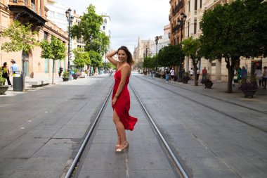 Kırmızı ipek elbiseli güzel bir kadın Seville 'de bir caddede tramvay raylarının arasında duruyor. Model fotoğraf çekimi için poz veriyor. Moda ve güzellik konsepti.