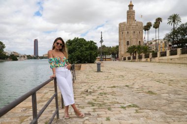 Young, pretty blonde woman with sunglasses leaning on the railing of the river. The woman is happy and smiling. In the background you can see the river in seville.