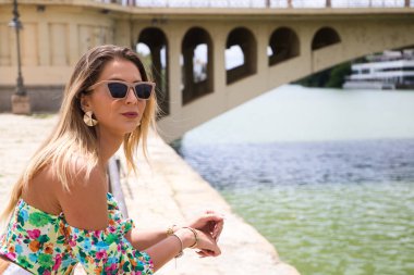 Pretty young blonde woman with sunglasses leaning over the railing of the river. The woman is looking at infinity while smiling or being serious. Various expressions.