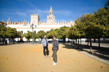 Young couple of gay men, walking in seville between orange trees, in the background you can see the gothic style cathedral and blue sky. The couple are married. Concept of gay rights