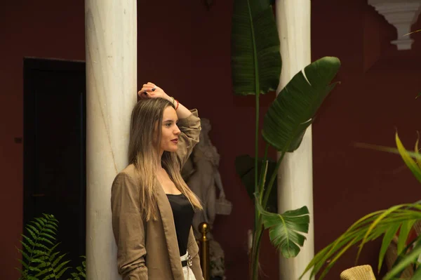 stock image Young and beautiful blonde Latin woman is leaning against a marble column in the inner courtyard of a hotel. The woman is sad and pensive with anguish. Concept depression and stress.